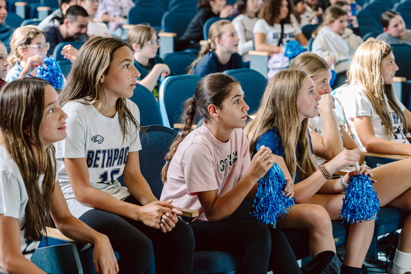 A group of people sitting in an auditorium, some holding blue pom-poms and backpacks.