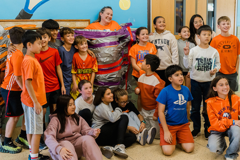 students sitting on floor in hall pose for a photo in front of person duct taped to wall