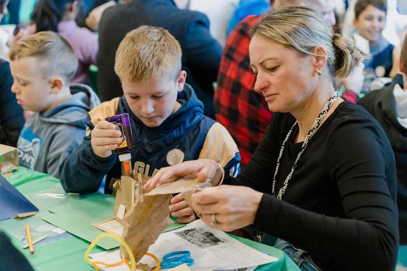 students are crafting at a table. They are focused on a paper project. The scene is lively, conveying creativity and concentration.