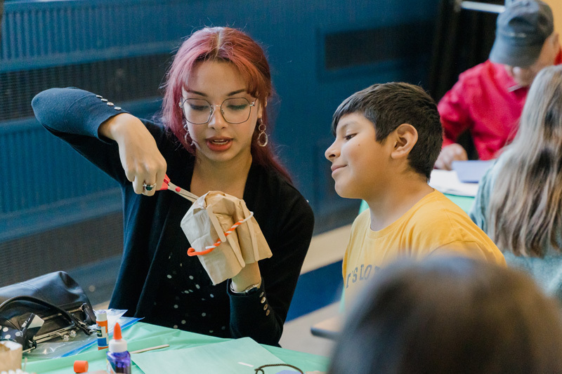 students are crafting at a table. They are focused on a paper project. The scene is lively, conveying creativity and concentration.