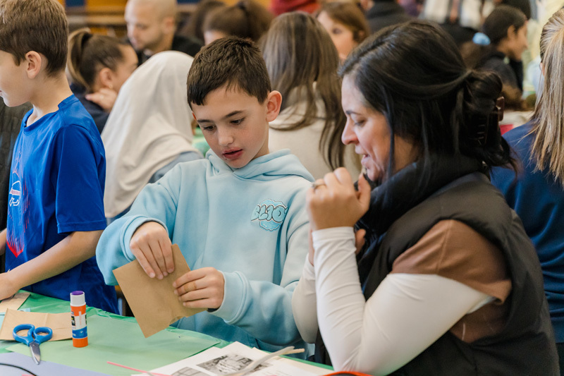 students are crafting at a table. They are focused on a paper project. The scene is lively, conveying creativity and concentration.