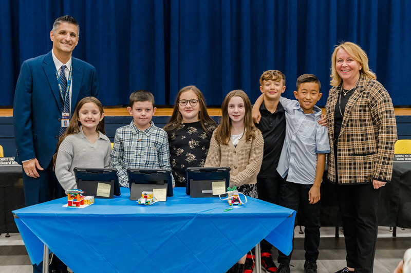 A group of six smiling children stands between two adults in a formal setting. 