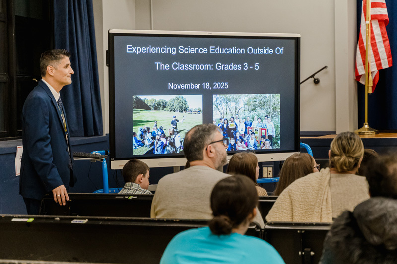 A presenter in a suit stands beside a projector screen displaying "Experiencing Science Education Outside of the Classroom: Grades 3-5," dated November 18, 2025.