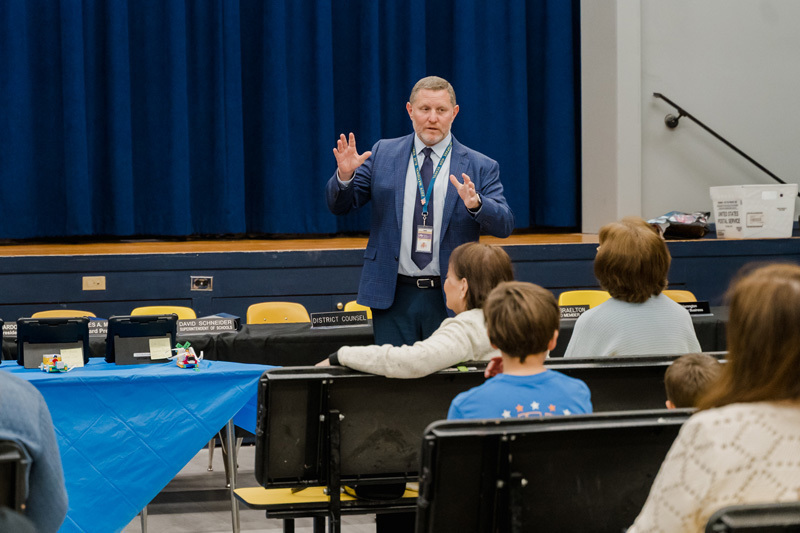 A man in a suit speaks to a small audience in a room with blue curtains. He gestures with his hands, conveying a sense of engagement and openness.
