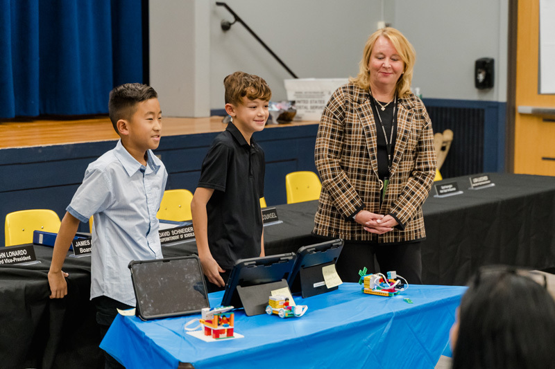 A group of smiling children stands up front of the class in a formal setting. 