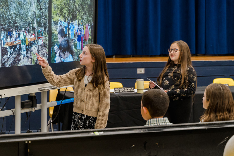students present confidently in front of an audience, standing by a screen 