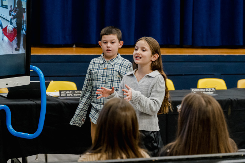 Two children present enthusiastically in front of a screen. They face an audience seated in a room with a blue curtain backdrop and yellow chairs.