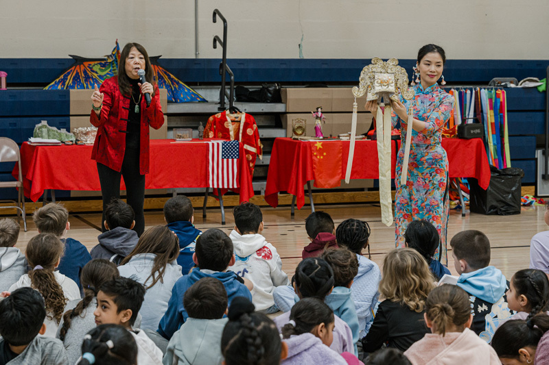 Two women present Chinese cultural items to seated children. One speaks into a microphone; the other holds a decorative headdress. Tables with flags, red cloths, and various objects are in the background. The mood is educational and engaging.