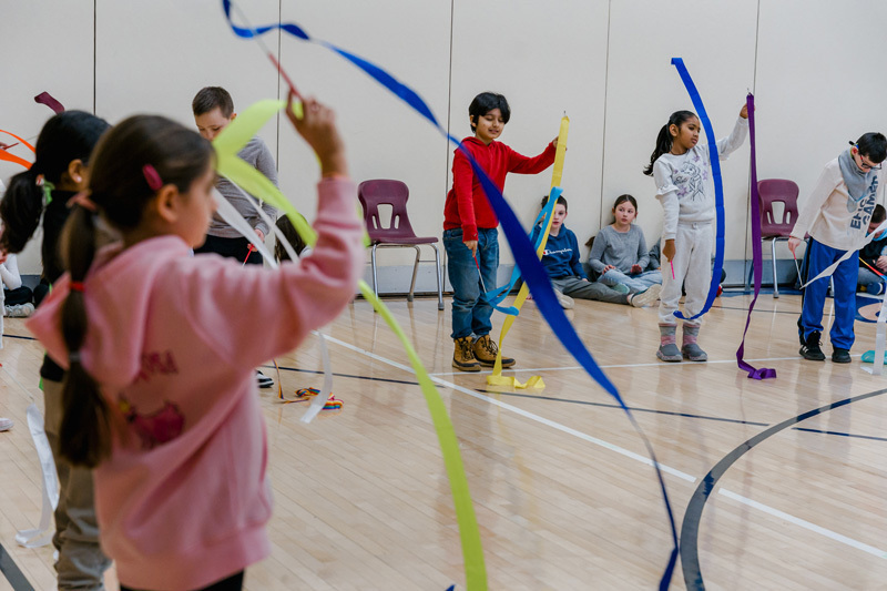 Children joyfully wave colorful ribbons in a gymnasium, creating a lively and playful atmosphere. Chairs and more children are visible in the background.