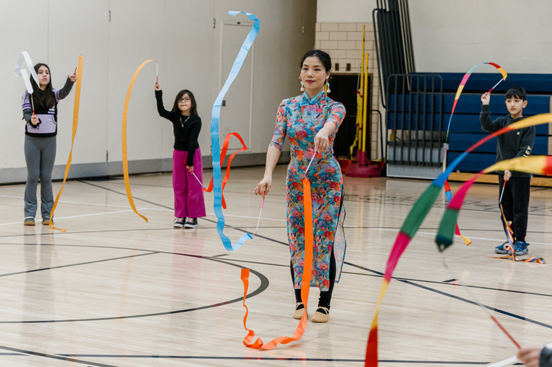 Children joyfully wave colorful ribbons in a gymnasium, creating a lively and playful atmosphere. Chairs and more children are visible in the background.