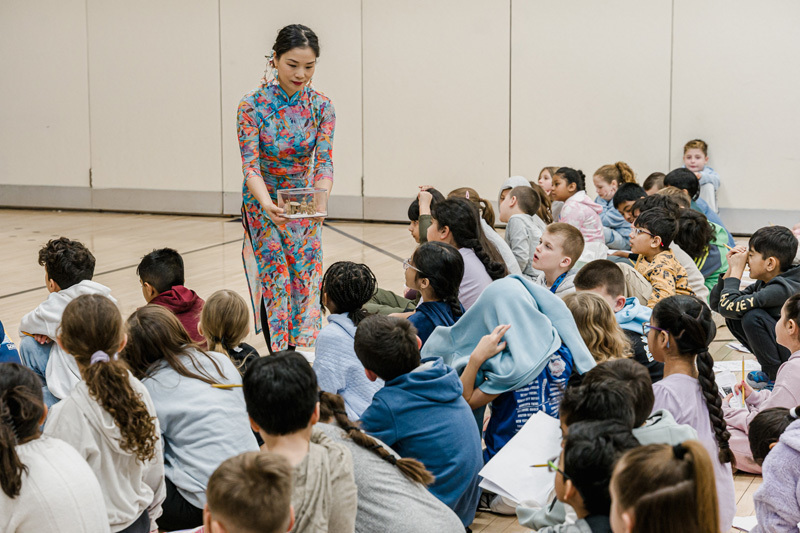Children joyfully wave colorful ribbons in a gymnasium, creating a lively and playful atmosphere. Chairs and more children are visible in the background.