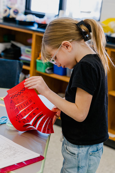 Children joyfully wave colorful ribbons in a gymnasium, creating a lively and playful atmosphere. Chairs and more children are visible in the background.