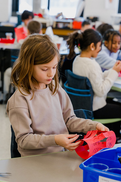 Children joyfully wave colorful ribbons in a gymnasium, creating a lively and playful atmosphere. Chairs and more children are visible in the background.