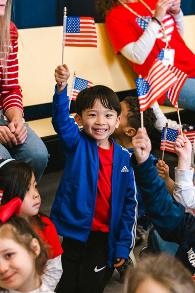 A diverse group of children sit closely together, some waving small American flags. A boy in a red shirt is smiling brightly, creating a joyful, patriotic atmosphere.