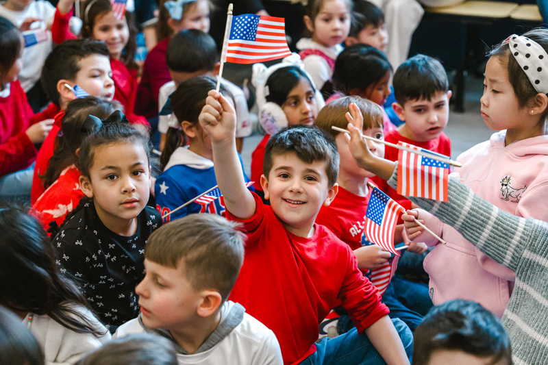 A diverse group of children sit closely together, some waving small American flags. A boy in a red shirt is smiling brightly, creating a joyful, patriotic atmosphere.