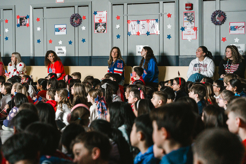 A diverse group of children sit closely together, some waving small American flags. A boy in a red shirt is smiling brightly, creating a joyful, patriotic atmosphere.