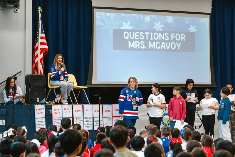 A diverse group of children sit closely together, some waving small American flags. A boy in a red shirt is smiling brightly, creating a joyful, patriotic atmosphere.