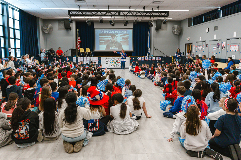 A diverse group of children sit closely together, some waving small American flags. A boy in a red shirt is smiling brightly, creating a joyful, patriotic atmosphere.