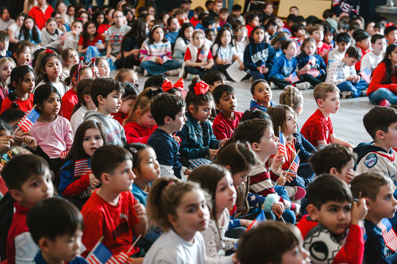 A diverse group of children sit closely together, some waving small American flags. A boy in a red shirt is smiling brightly, creating a joyful, patriotic atmosphere.