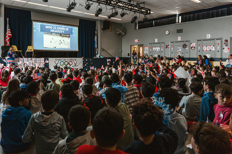A diverse group of children sit closely together, some waving small American flags. A boy in a red shirt is smiling brightly, creating a joyful, patriotic atmosphere.