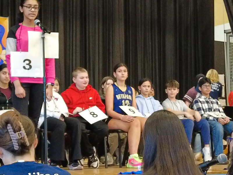 A group of students sit on a stage for a spelling bee, each with a numbered card. A banner overhead reads "4th Annual Schoolwide Spelling Bee." The audience watches attentively.