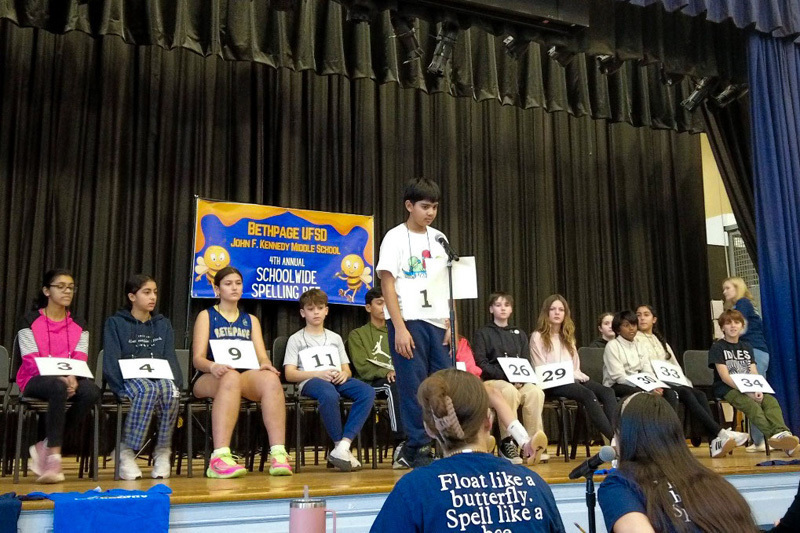 A group of students sit on a stage for a spelling bee, each with a numbered card. A banner overhead reads "4th Annual Schoolwide Spelling Bee." The audience watches attentively.