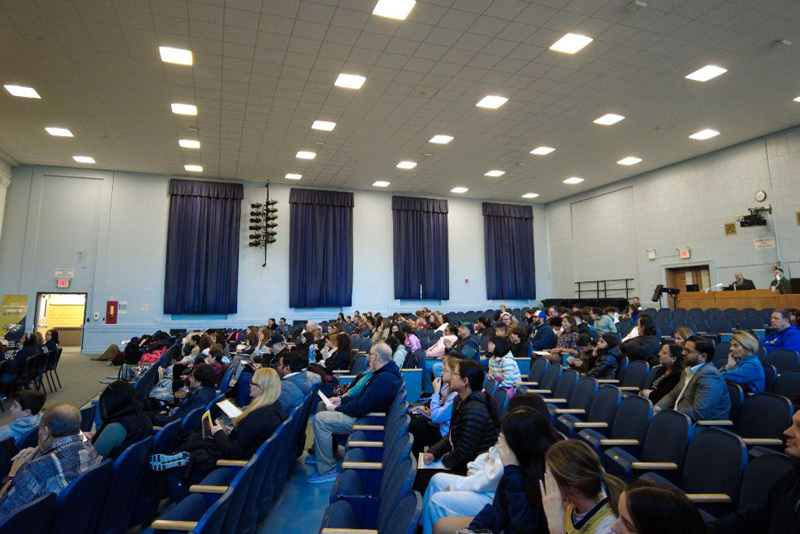 A group of students sit on a stage for a spelling bee, each with a numbered card. A banner overhead reads "4th Annual Schoolwide Spelling Bee." The audience watches attentively.