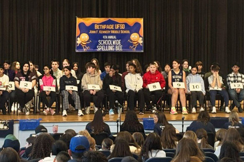 A group of students sit on a stage for a spelling bee, each with a numbered card. A banner overhead reads "4th Annual Schoolwide Spelling Bee." The audience watches attentively.