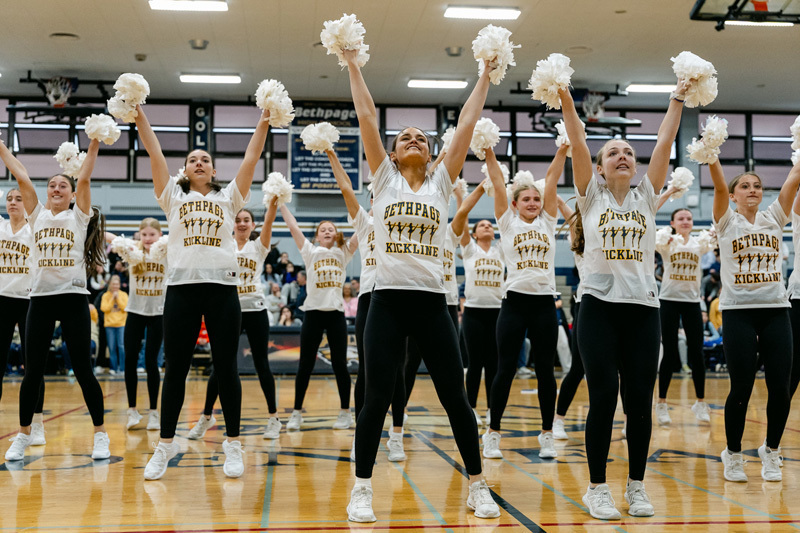 A group of cheerleaders in matching uniforms enthusiastically raise pom-poms in a gymnasium. The atmosphere is energetic and focused.