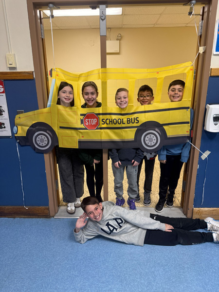 A diverse group of smiling children and two adults pose in a classroom. In the background, a yellow “school bus” decoration adds a playful vibe.
