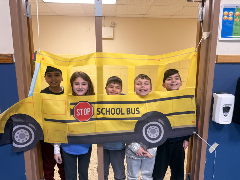 A diverse group of smiling children and two adults pose in a classroom. In the background, a yellow “school bus” decoration adds a playful vibe.
