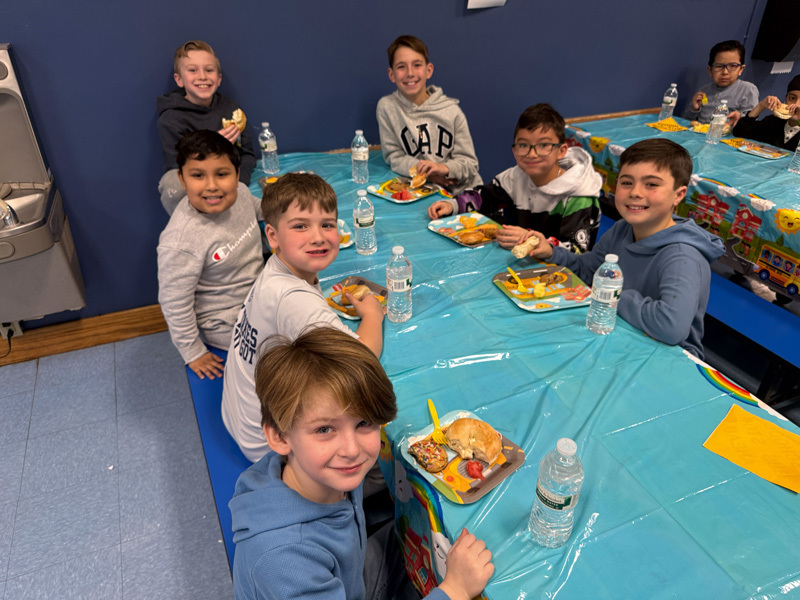 A diverse group of smiling children and two adults pose in a classroom. In the background, a yellow “school bus” decoration adds a playful vibe.