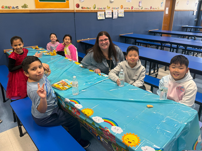 A diverse group of smiling children and two adults pose in a classroom. In the background, a yellow “school bus” decoration adds a playful vibe.
