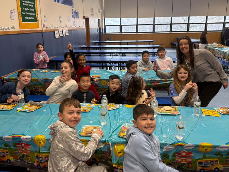 A diverse group of smiling children and two adults pose in a classroom. In the background, a yellow “school bus” decoration adds a playful vibe.
