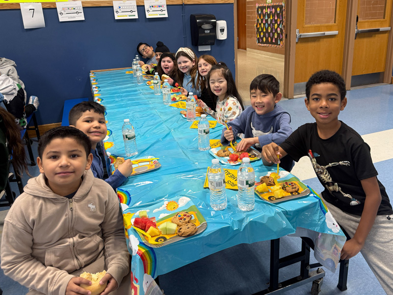 A diverse group of smiling children and two adults pose in a classroom. In the background, a yellow “school bus” decoration adds a playful vibe.