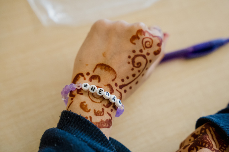 A hand adorned with intricate henna designs and a bracelet made of white and purple beads, reading "NERA." The background is a light wooden surface.