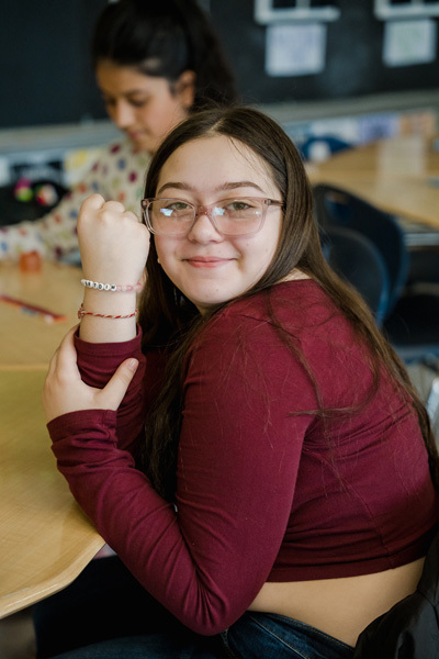 student shows off a beaded bracelet, sitting at a classroom table. 