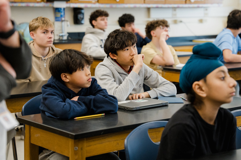 A group of students sits attentively at lab tables in a classroom, 
