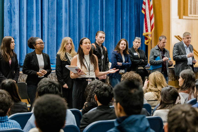 A diverse group of professionals, dressed in business attire, stand on stage