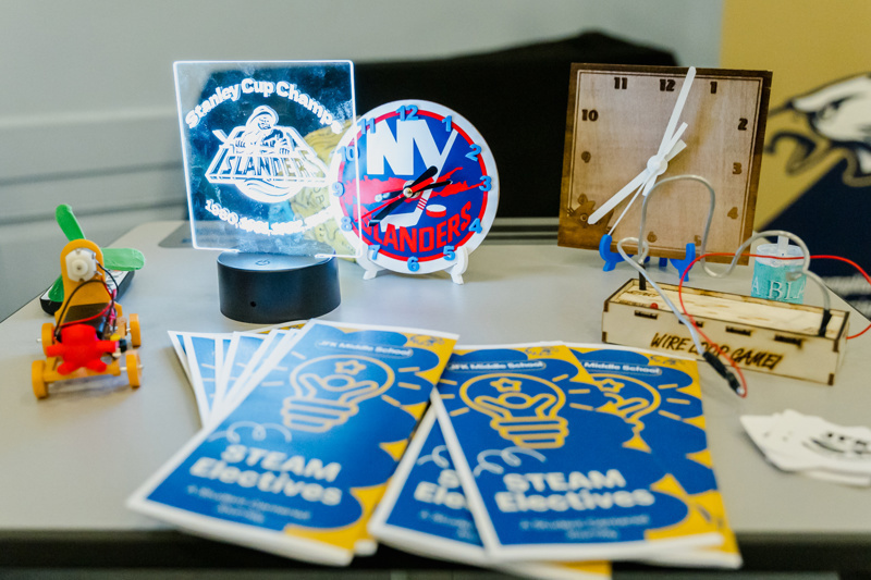 A tabletop display featuring a Stanley Cup Champions plaque, a colorful New York Islanders clock, educational STEAM elective brochures, a wooden clock model, and a small toy airplane.