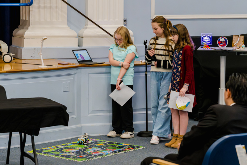 Students presenting a robotics project at a school event, showcasing teamwork and creativity while demonstrating their robot on a colorful play mat.