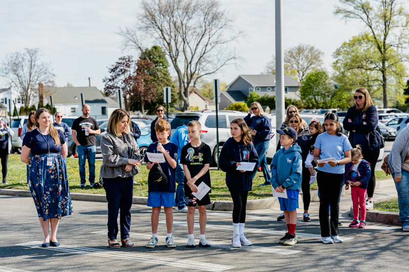 Students and adults participate in a community event, standing together in a parking lot, with an audience in the background.