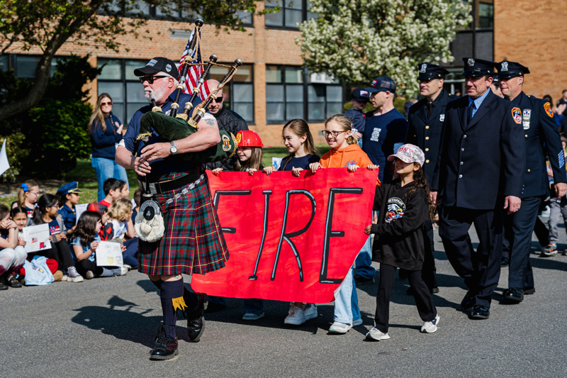 Children participating in a community event hold a large red banner reading "FIRE," while firefighters and other participants gather in the background.