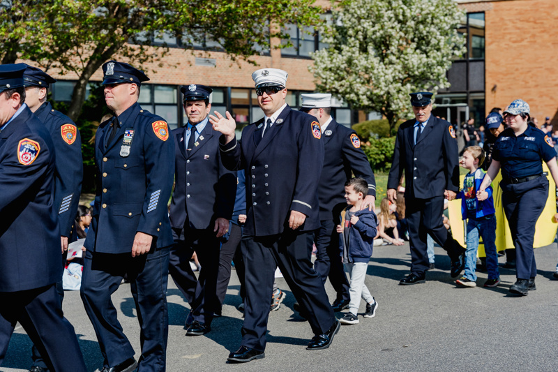 First responders in uniform marching in a parade, with children participating alongside them in a community celebration.
