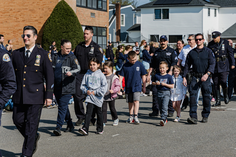 First responders in uniform marching in a parade, with children participating alongside them in a community celebration.