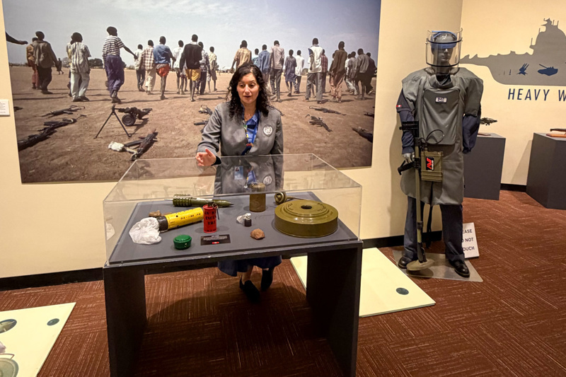 A woman presents historical artifacts related to conflict resolution at an exhibit, featuring a display case with various items and a background of individuals walking in a dry landscape, highlighting themes of peace and war.