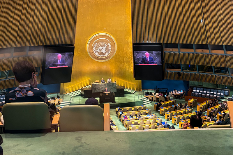 United Nations General Assembly session in progress, featuring speakers at the podium with audience members and large screens displaying the event.