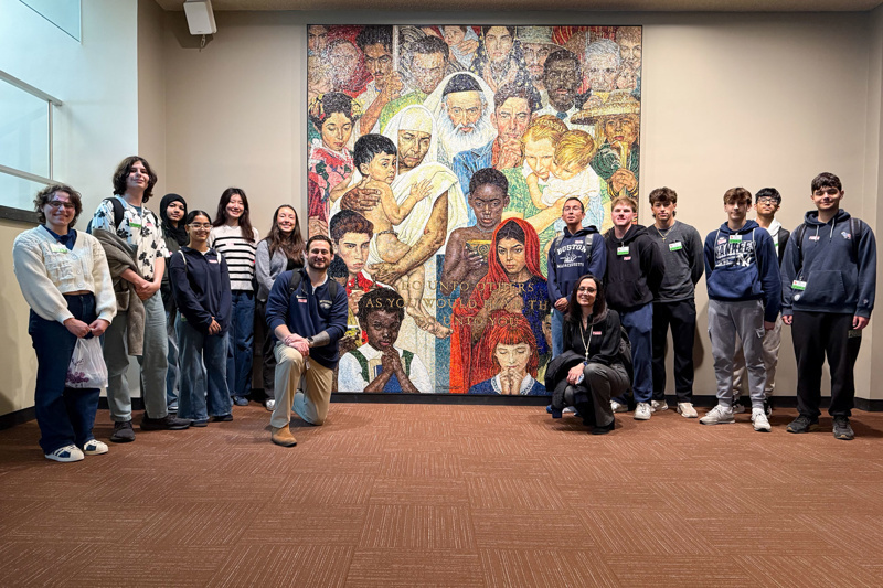 Group of students and educators posing in front of a colorful mural depicting diverse individuals, highlighting themes of community and support.