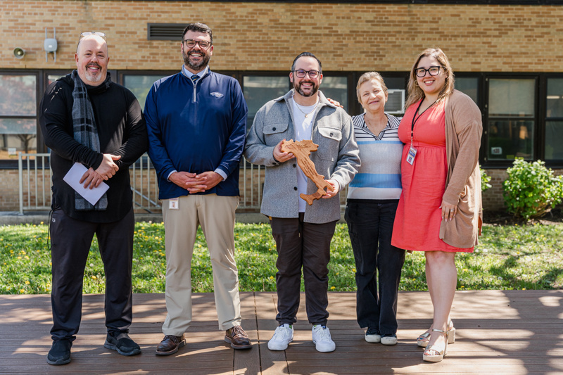 Group photo of five individuals standing outdoors, smiling together, with one person holding a wooden award. The setting highlights a sunny day near a school building, showcasing community spirit and celebration.