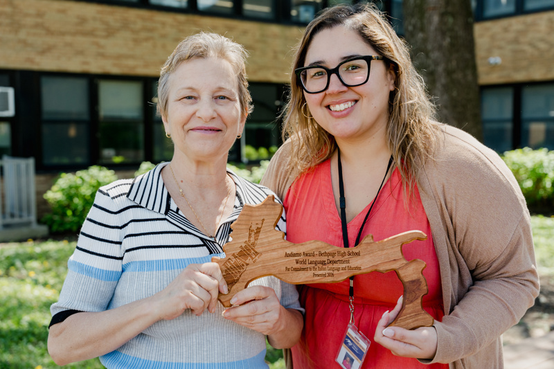 Two women proudly hold a wooden award outside a school, celebrating achievements in the World Language Department at Bethpage High School.